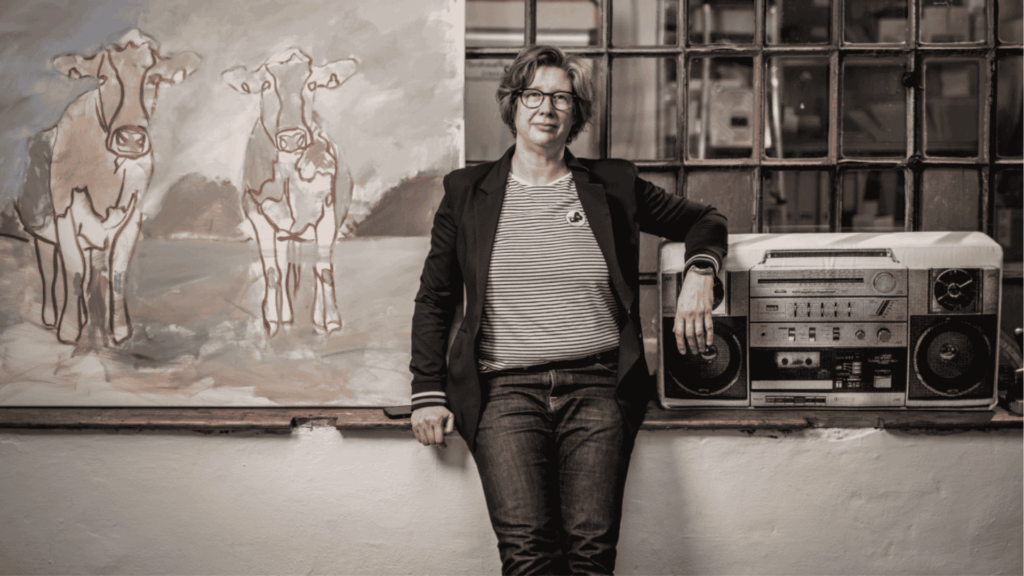 Maren Martschenko steht und sitzt leicht angelehnt an einem Absatz. Neben ihr ein Ghettoblaster und hinter ihr hängt eine Weltkarte.