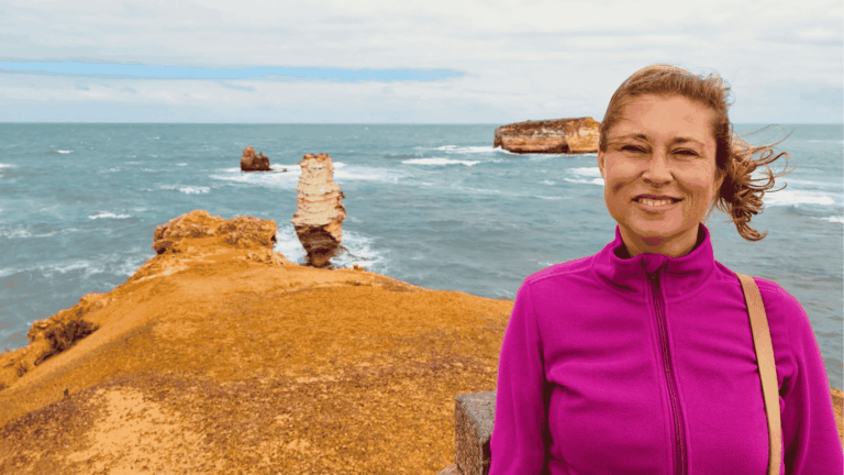 Claudia Sittner von Modern Sabbatical trägt eine pinkfarbene Jacke und lächelt in die Kamera. Sie steht draußen und hinter ihr erkennt man das Meer.