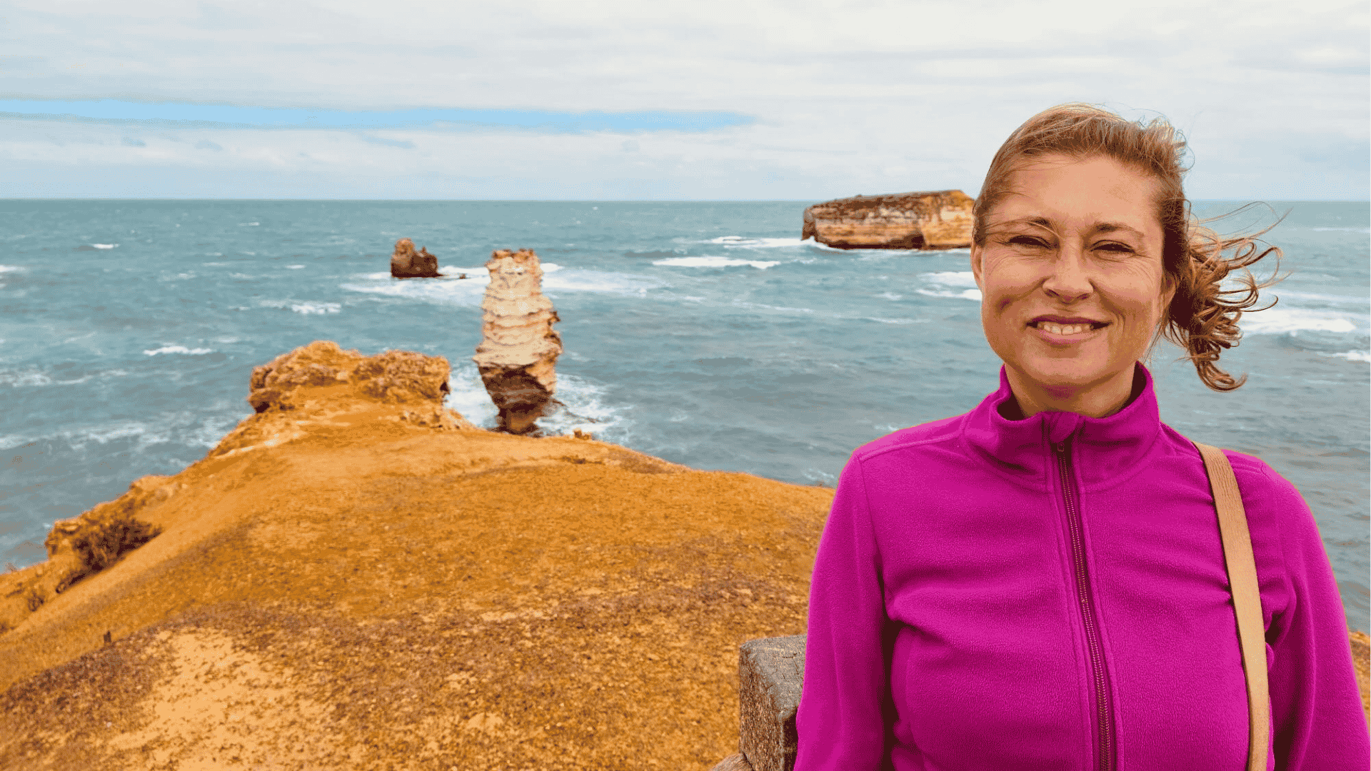 Claudia Sittner von Modern Sabbatical trägt eine pinkfarbene Jacke und lächelt in die Kamera. Sie steht draußen und hinter ihr erkennt man das Meer.
