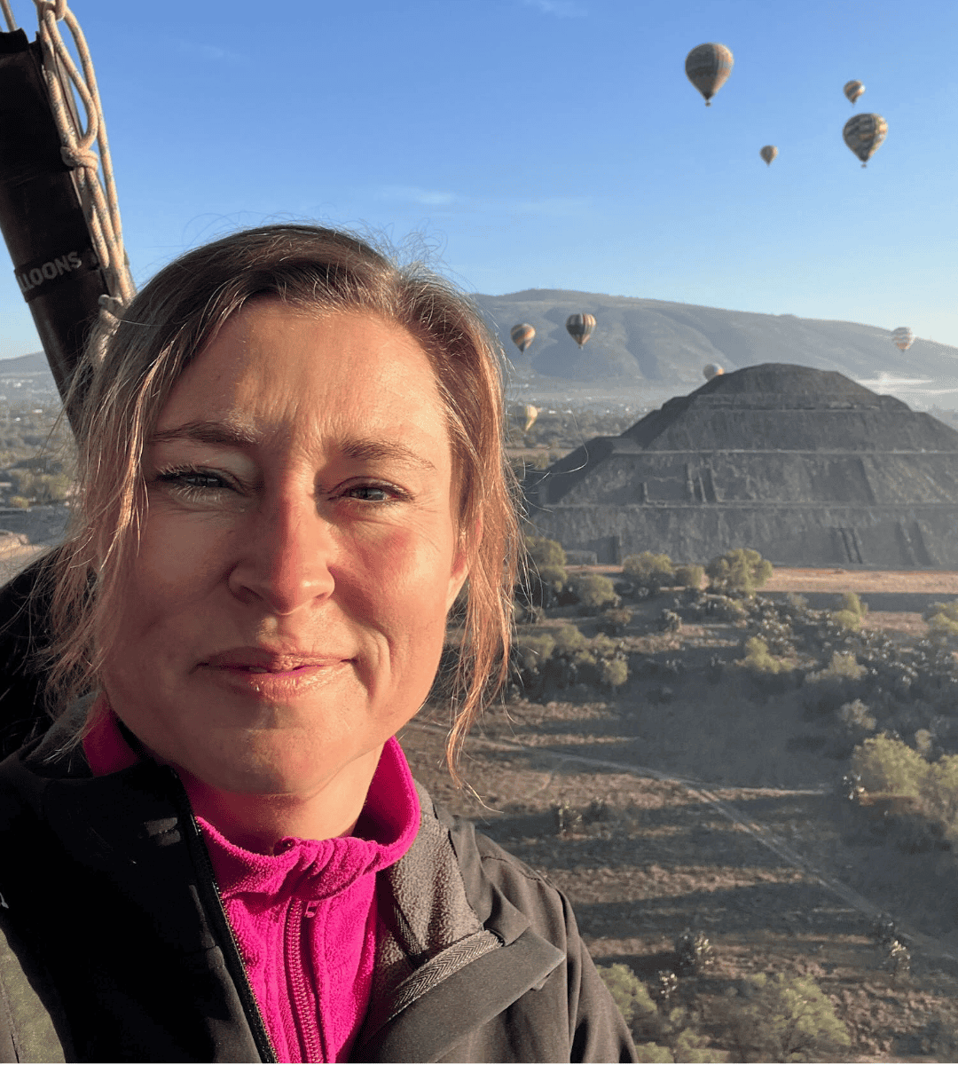 Claudia in einem Heißluftballon in Mexiko. Sonne leuchtet ihr Gesicht aus und hinter ihr fahren viele weitere Ballons in der Ferne.